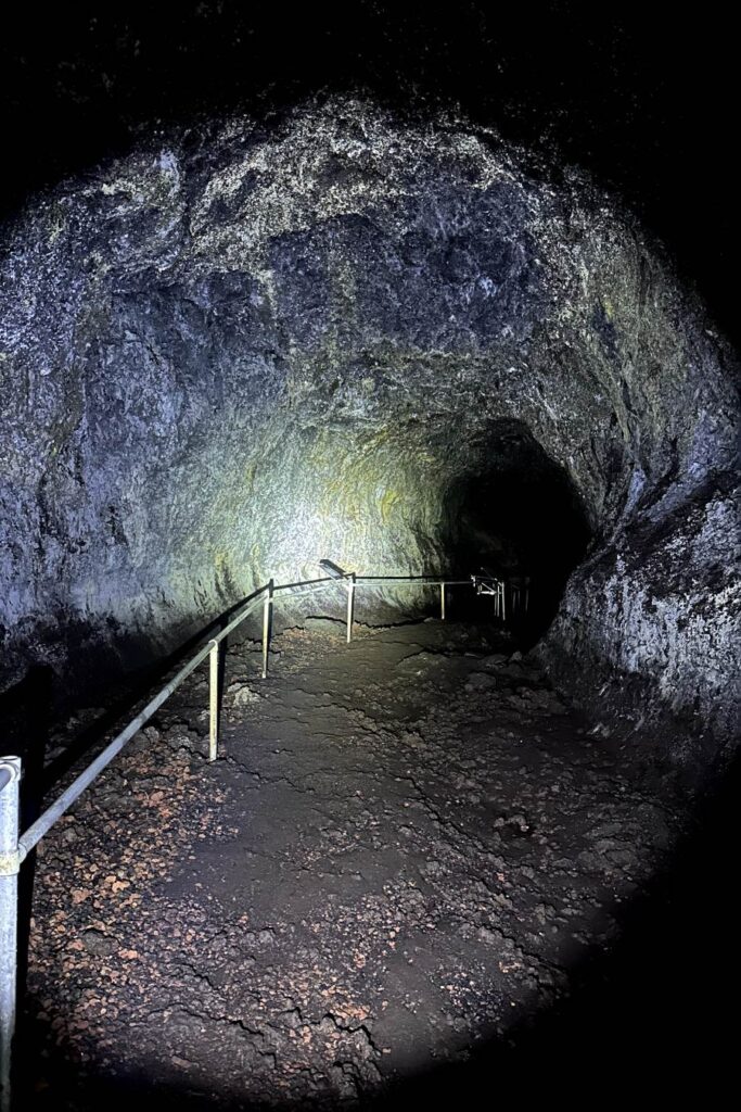 A dark passage inside the Hana Lava Tube with rocky walls illuminated by a flashlight, showing a winding path with metal handrails. Must do in Maui