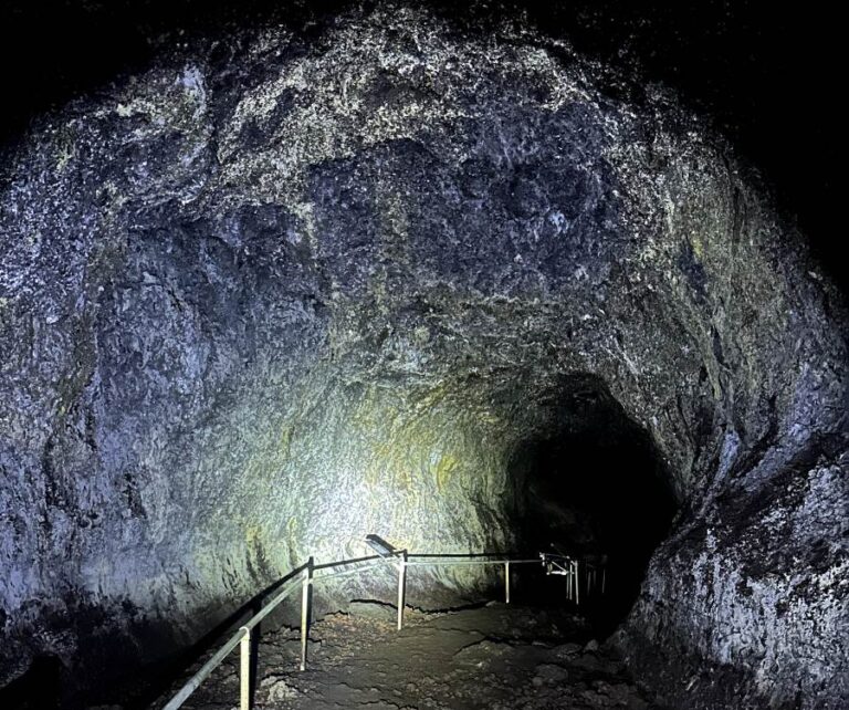 A dark passage inside the Hana Lava Tube with rocky walls illuminated by a flashlight, showing a winding path with metal handrails.