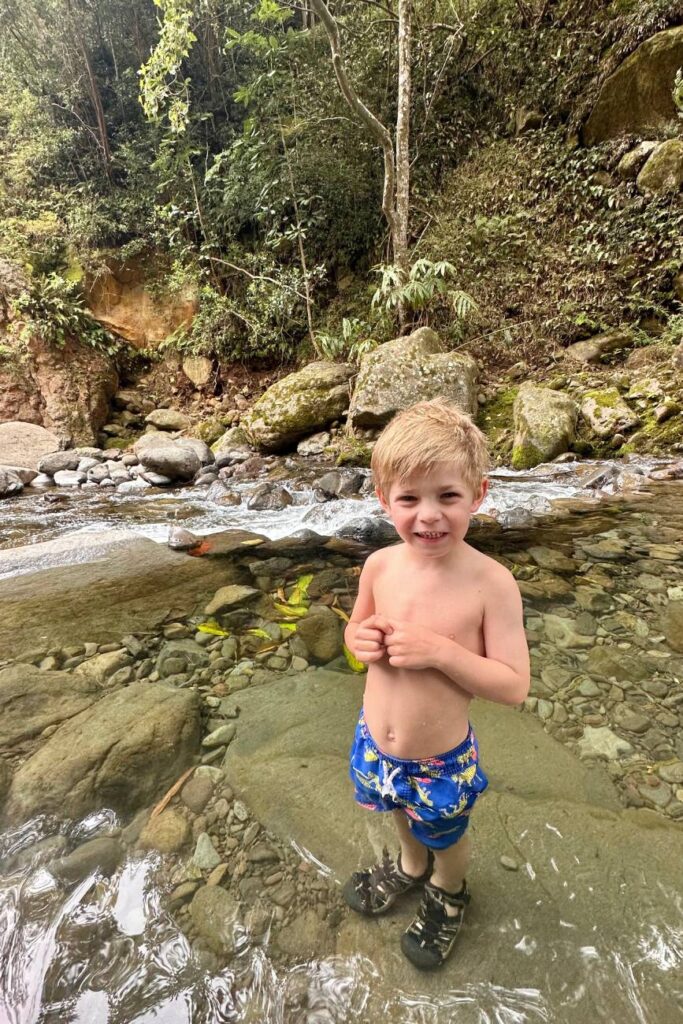 A boy stands smiling in a shallow, clear stream surrounded by rocks and forest in ʻĪao Valley. must do in Maui
