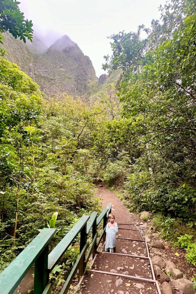 A young child wrapped in a towel stands on a dirt trail with stairs in the lush forest of ʻĪao Valley.