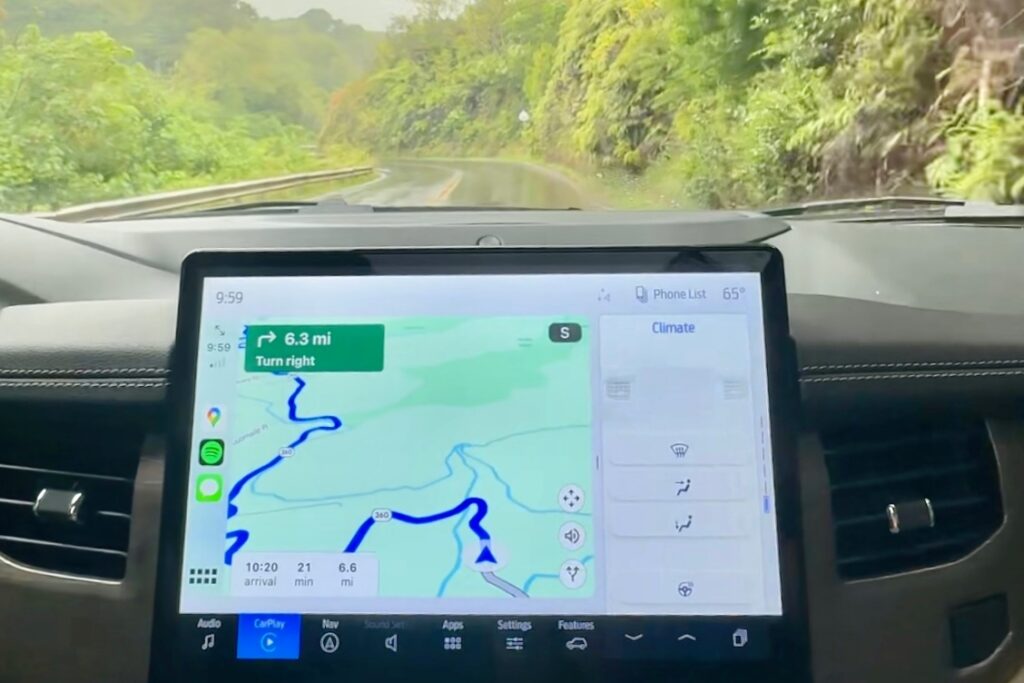 Car navigation screen showing winding road directions on the Road to Hana, with lush green landscape visible through the windshield. Maui, Hawaii.