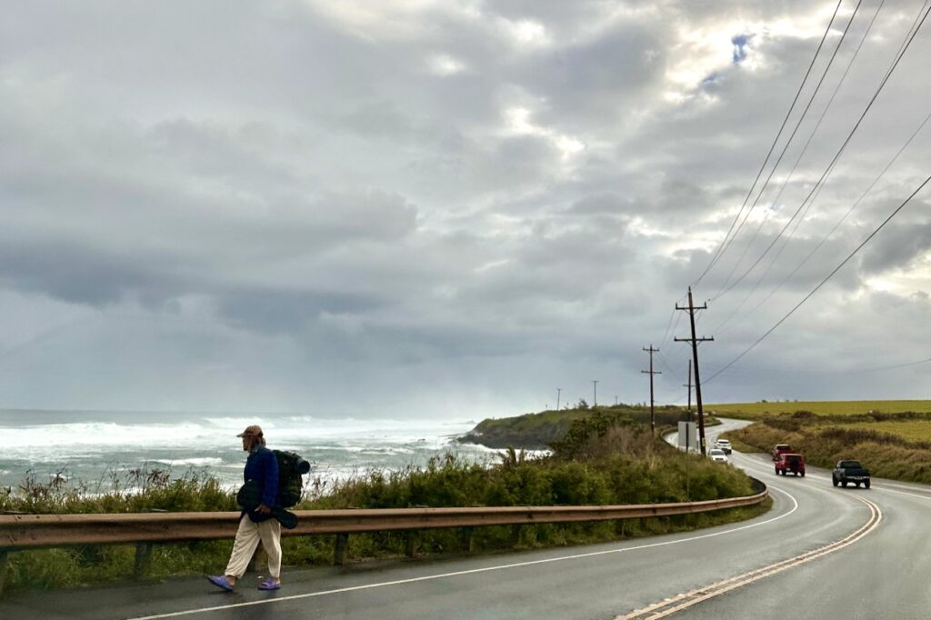 Scenic coastal road on a cloudy day with cars driving alongside crashing ocean waves, bordered by green fields and power lines. Maui, Hawaii.