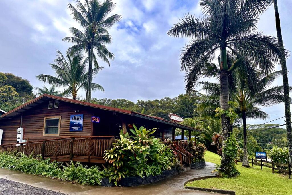Rustic wooden building with a sign reading "Jaws Country Store," surrounded by palm trees and tropical plants along the Road to Hana. Maui, Hawaii.