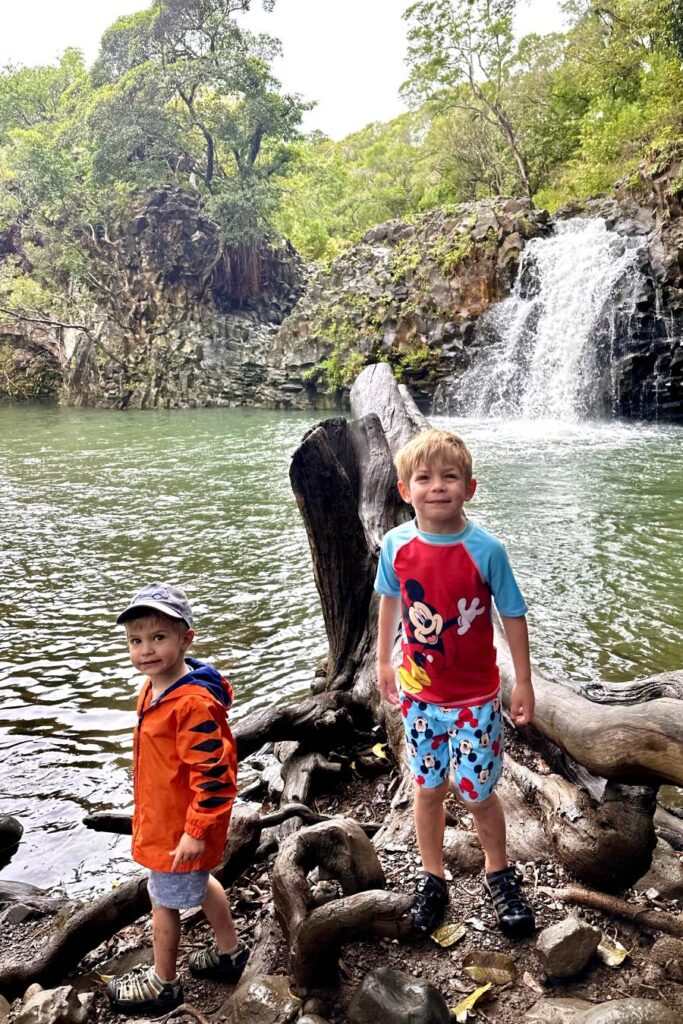 Two young boys in swimwear standing near a waterfall in a lush, natural setting with green water and surrounding rock formations. Maui, Hawaii.