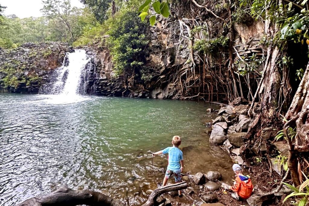 Young children playing near a calm green pool with a waterfall, surrounded by large rocks, tree roots, and lush vegetation. Maui, Hawaii.