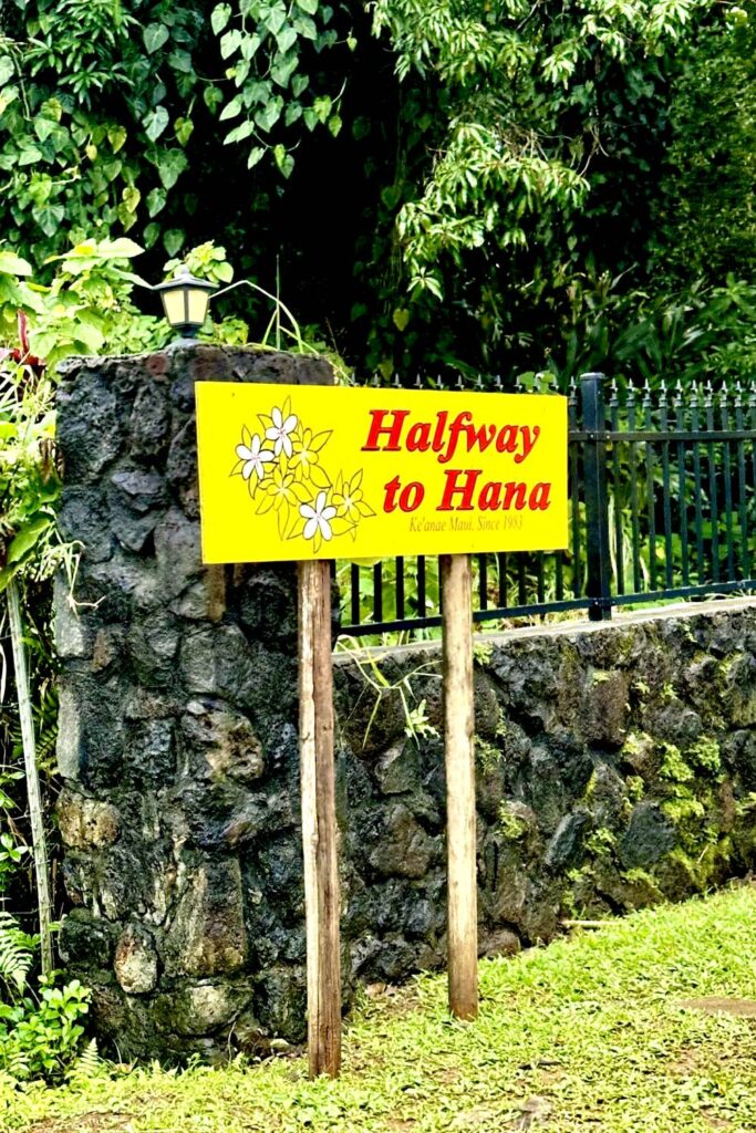 A bright yellow sign that reads "Halfway to Hana," mounted on a rustic stone wall with tropical plants in the background. Maui, Hawaii.