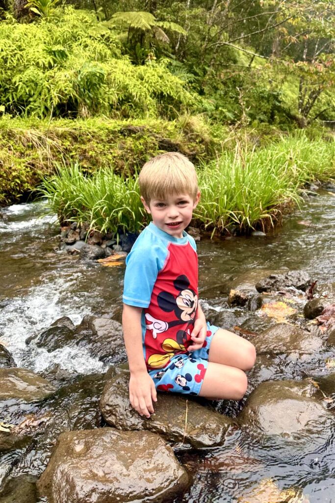 A young child in a Mickey Mouse swimsuit sits on rocks by a gentle stream, smiling and surrounded by lush greenery and flowing water. Maui, Hawaii.