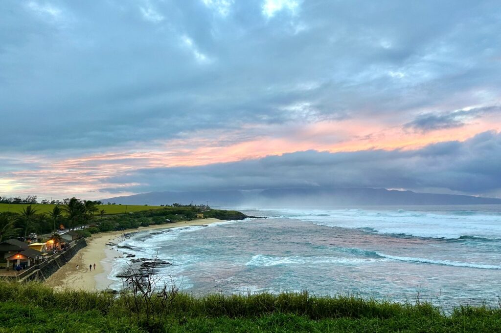 A beautiful beach scene at sunset with a small group of people walking along the shore, palm trees, and gentle waves with colorful clouds in the sky above. Maui, Hawaii.