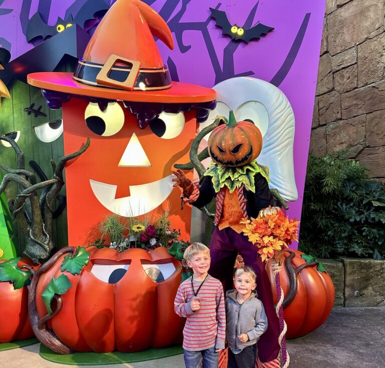 Two young boys stand with a person on stilts with a jack-o-lantern head in front of a Halloween display