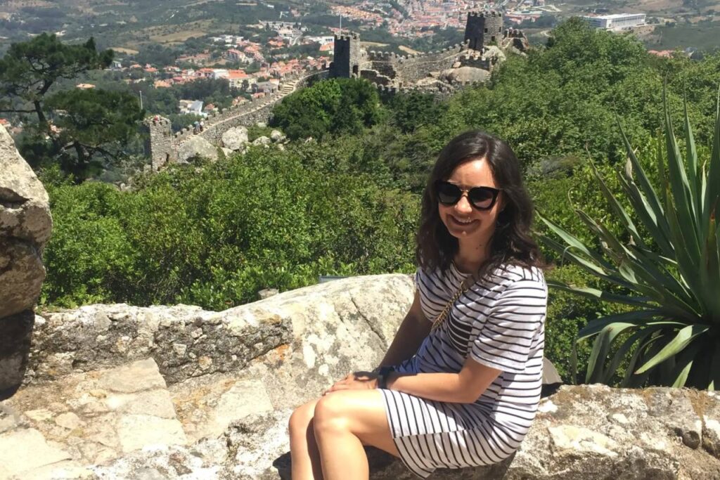 Woman sitting on a stone wall at the Castle of the Moors in Sintra, Portugal, with fortress walls and green hills behind her.
