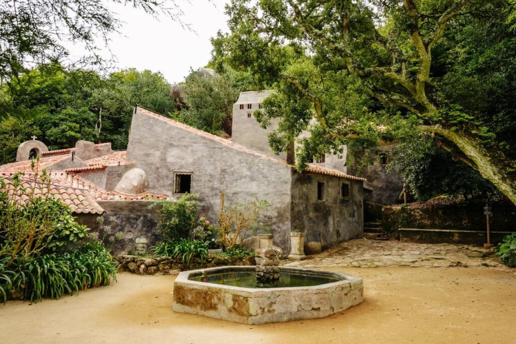 Convent of the Capuchos in Sintra, Portugal, with simple stone buildings, terracotta rooftops, and a small fountain surrounded by trees and greenery.