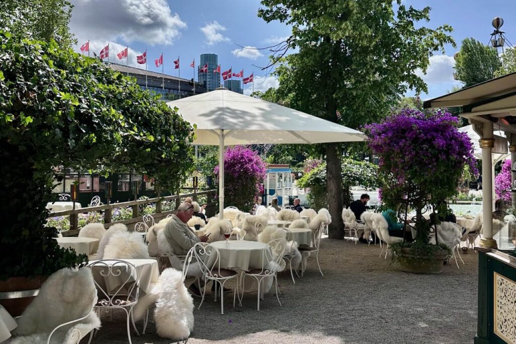 Outdoor café at Tivoli Gardens with white wrought-iron chairs covered in fluffy sheepskin throws, shaded by umbrellas and surrounded by flowers.