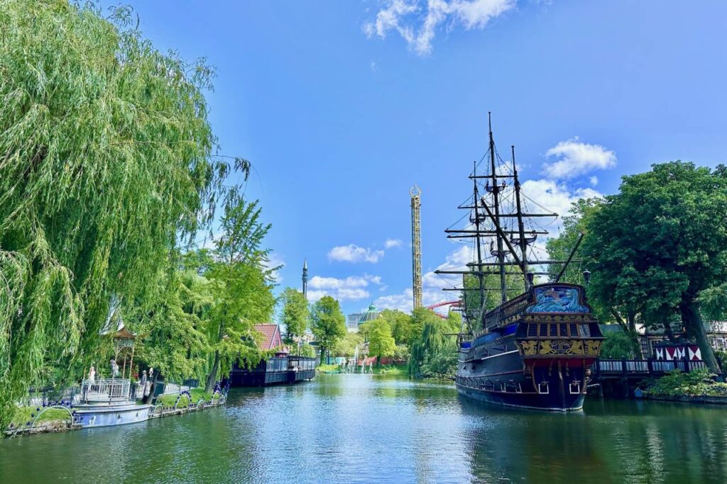 Scenic view of Tivoli Gardens’ lake with lush greenery, boats, and amusement rides in the distance.