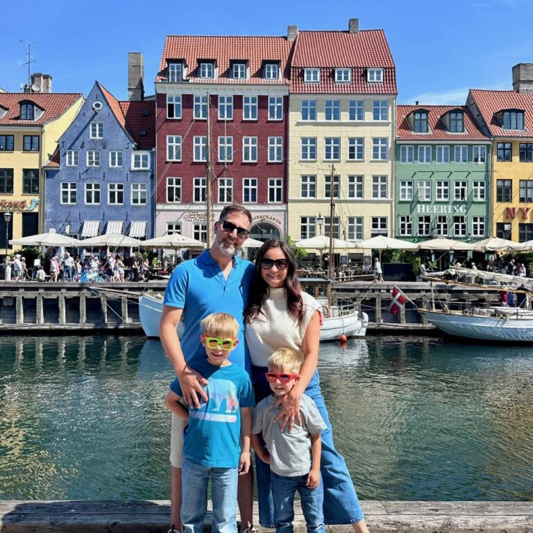 A family of four smiles in front of the colorful townhouses along Nyhavn canal on a sunny day during a Copenhagen itinerary.