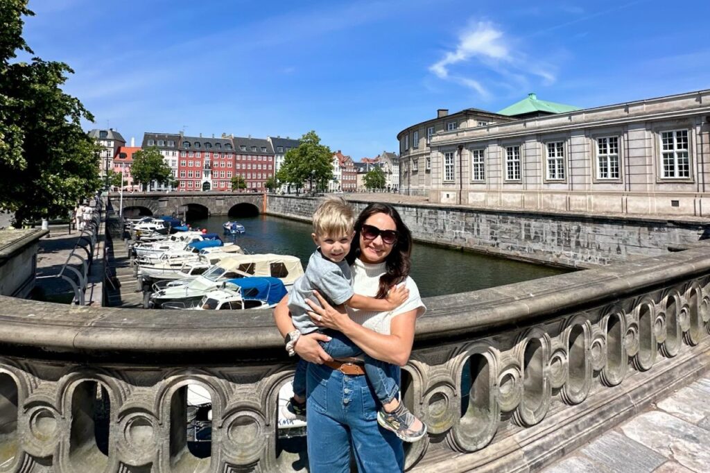 A mom holds her young son while standing on a bridge overlooking a Copenhagen canal filled with small boats.