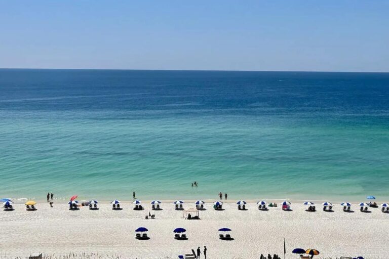 Aerial view of Pensacola Beach with rows of blue and white umbrellas lining the soft, white sand and people enjoying the clear, calm turquoise waters of the Gulf of Mexico.