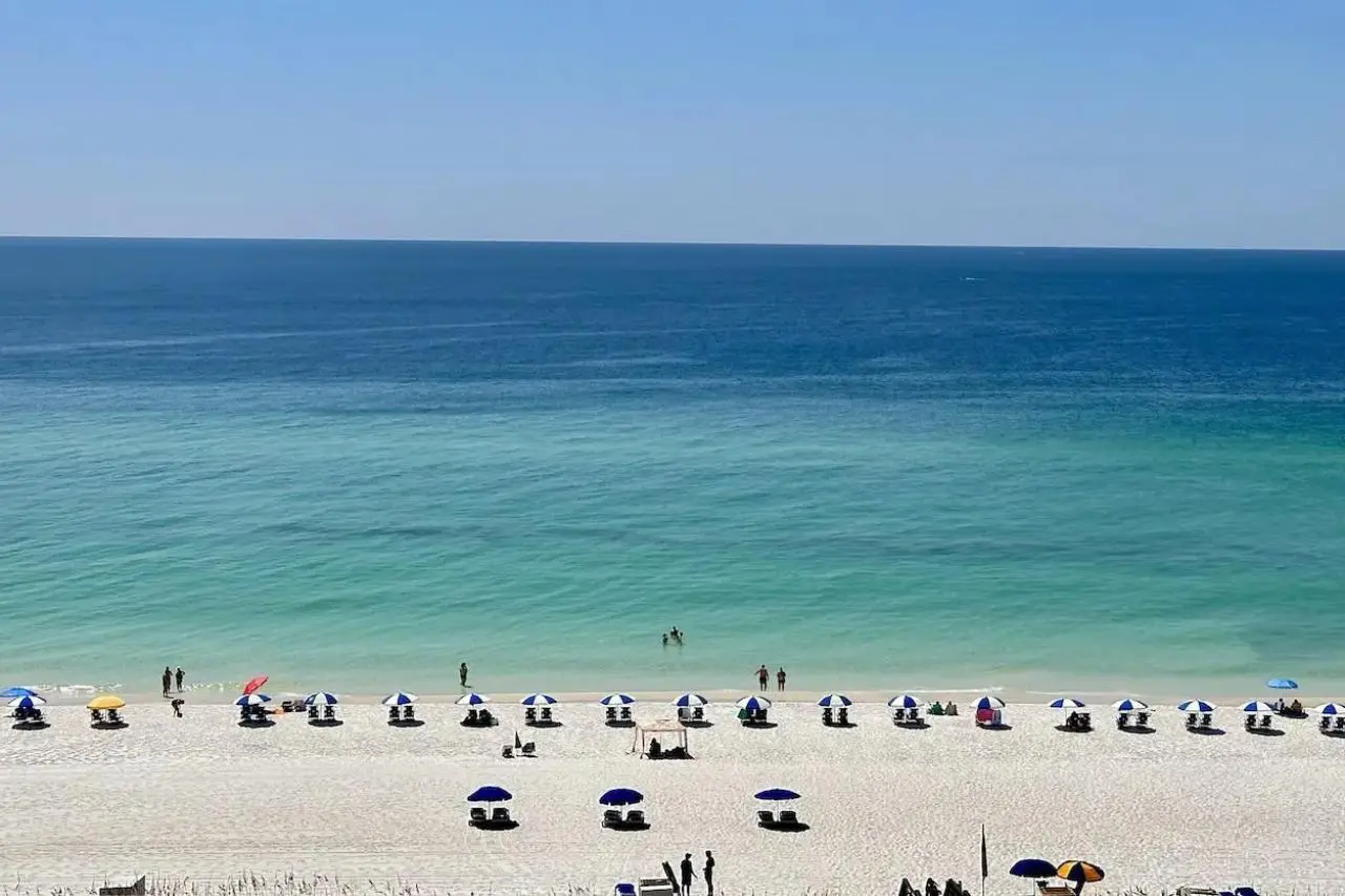 Aerial view of Pensacola Beach with rows of blue and white umbrellas lining the soft, white sand and people enjoying the clear, calm turquoise waters of the Gulf of Mexico.