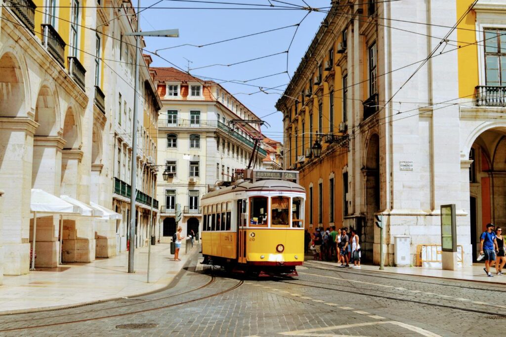 Yellow Lisbon tram rolling along tracks near Praça do Comércio, with pastel buildings, overhead wires, and pedestrians on a sunny day. Best countries for family travel