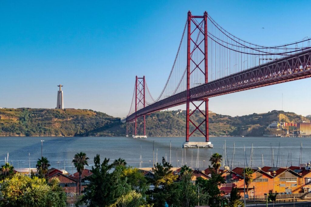 The red 25 de Abril Bridge stretching over the Tagus River, with the Cristo Rei statue on the hill and boat masts in the foreground.