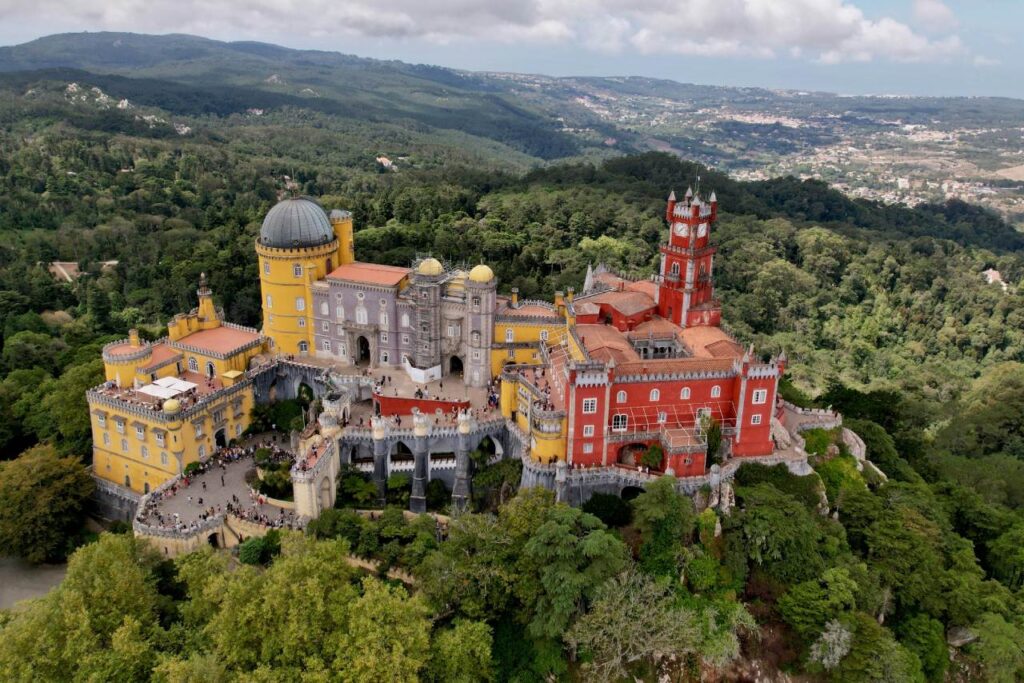 Aerial view of Pena Palace in Sintra, showing the colorful yellow and red towers surrounded by green forested hills.
