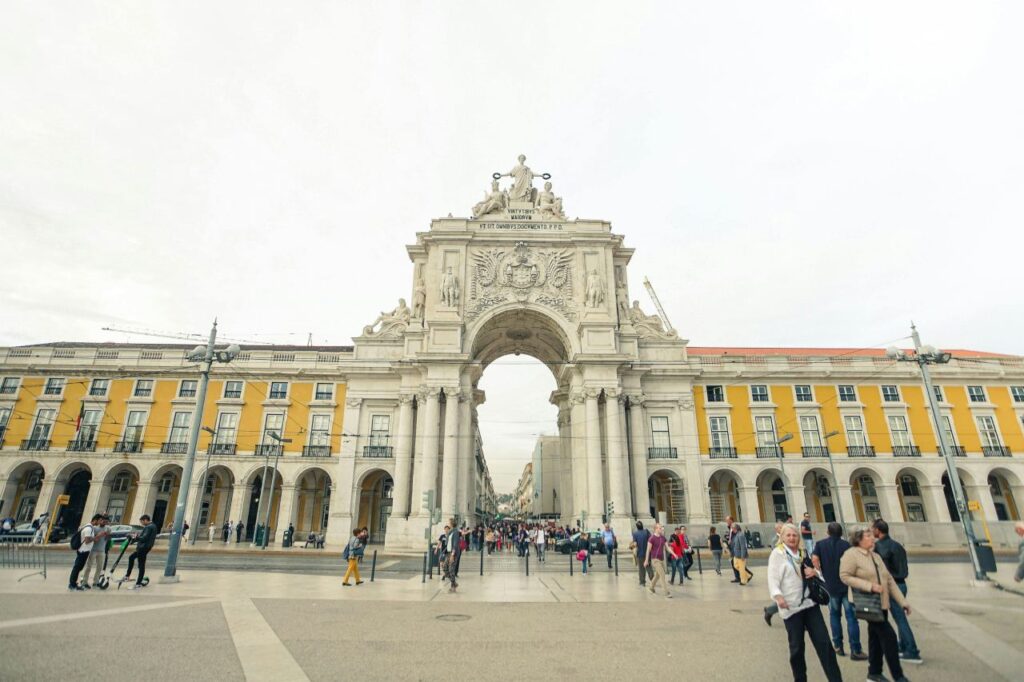 Arco da Rua Augusta at Praça do Comércio in Lisbon, with crowds walking across the square and yellow arcaded buildings.