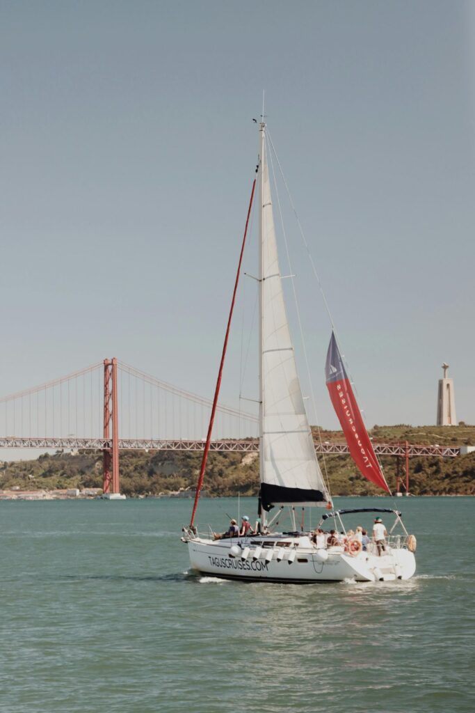 Sailboat cruising on the Tagus River with the 25 de Abril Bridge and Cristo Rei statue in the background.