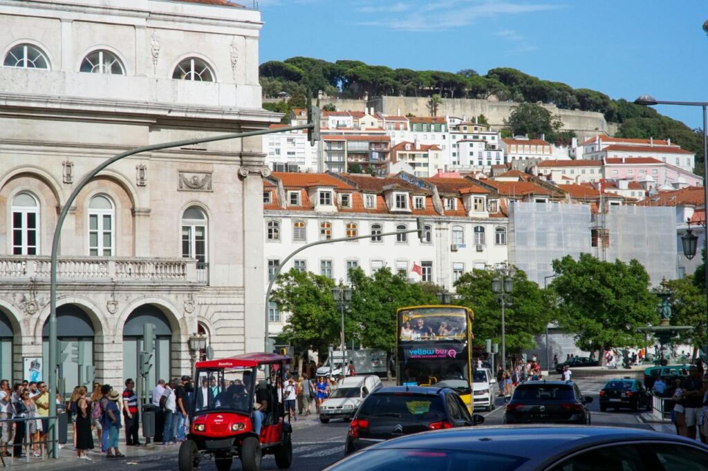 Lisbon street scene with a hop on hop off bus and a red tuk tuk in traffic, with historic buildings and a hillside backdrop.