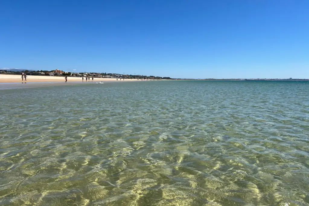 Shallow, clear water over sand on the wide beach of Meia Praia in Lagos, with people walking along the shoreline in the distance.