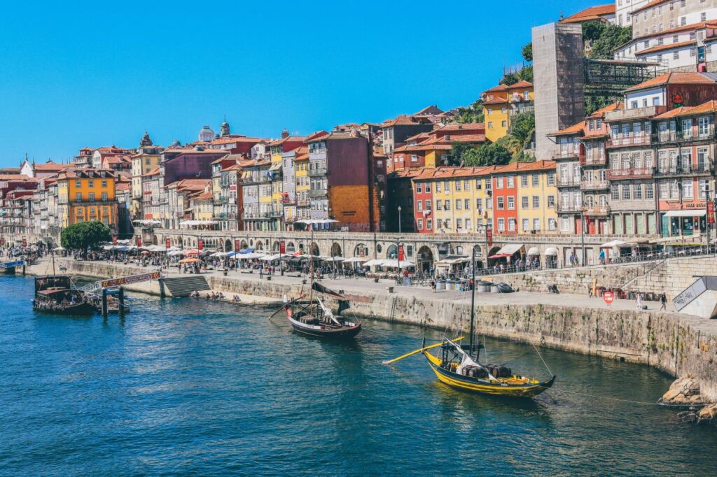 Colorful houses stacked along the Ribeira waterfront in Porto, Portugal, with traditional rabelo boats on the Douro River on a sunny day