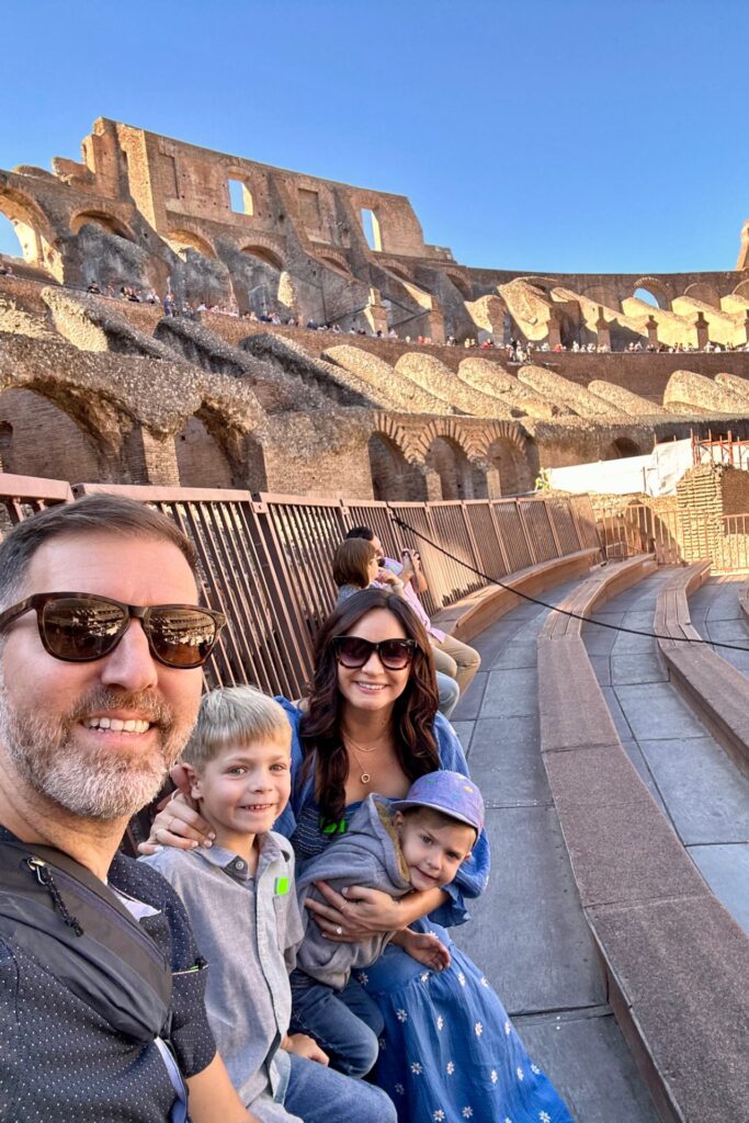Family selfie inside the Colosseum in Rome, with parents and two young boys seated in the ancient amphitheater.