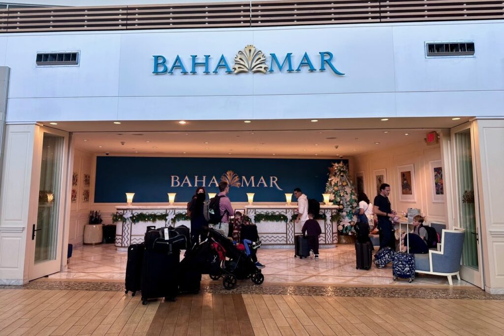 Families checking in at the Baha Mar resort lobby in Nassau, Bahamas, with luggage and strollers in the foreground and a Christmas tree visible at the front desk.
