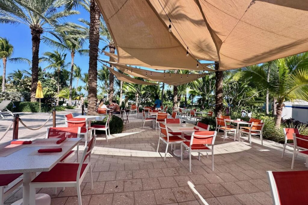 Outdoor dining terrace at Baha Mar shaded by large canvas sails, with red and white chairs, string lights, and palm trees framing a view of the beach.