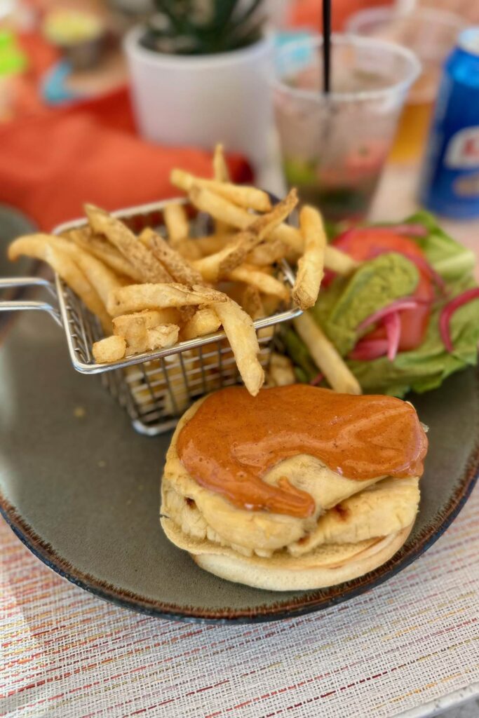 A sauced chicken sandwich served with a basket of thin-cut fries and fresh toppings on a dark plate at a Baha Mar restaurant.