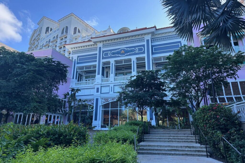 Street-level view of a colorful Baha Mar building with blue and white colonial-style architecture, lush tropical greenery, and the Grand Hyatt tower visible in the background.