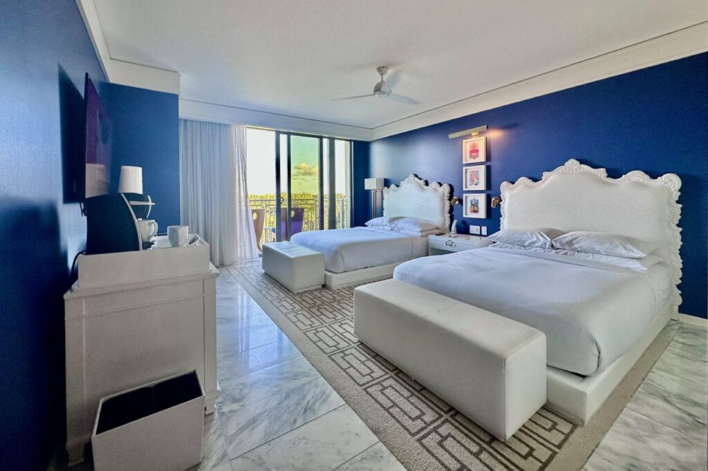 Renovated Grand Hyatt Baha Mar guest room with two queen beds, deep blue accent walls, ornate white headboards, marble floors, and a balcony with a view of the resort grounds.