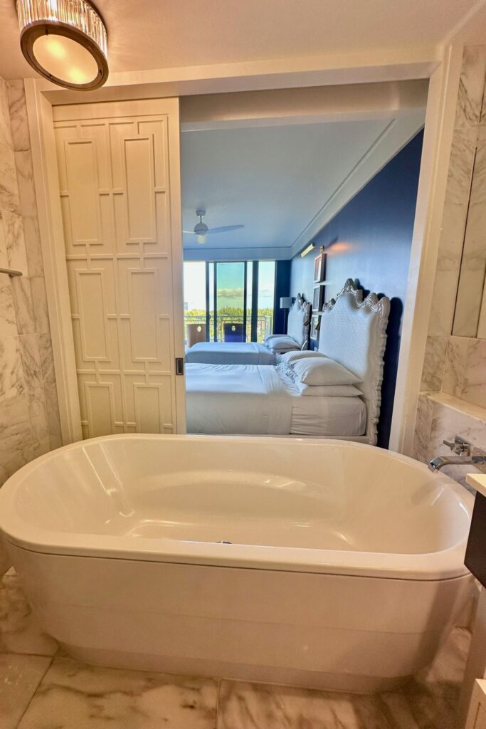 Freestanding soaking tub in the marble bathroom of a Grand Hyatt Baha Mar room, with a sliding door opening to the bedroom beyond.