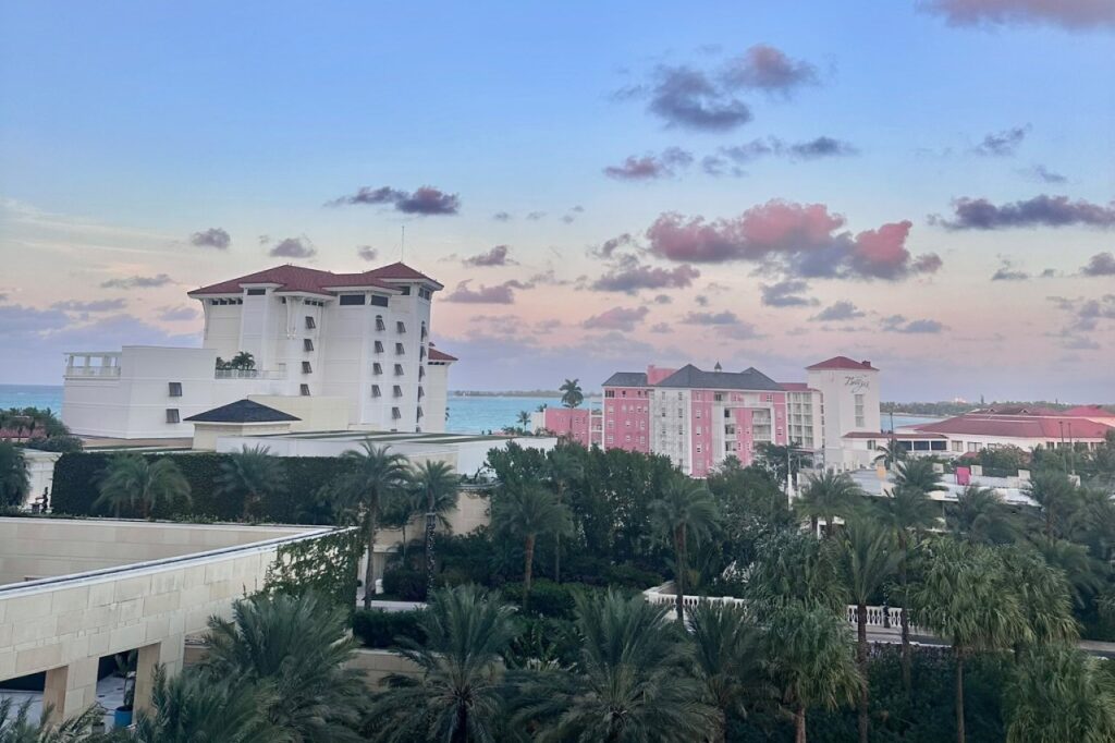 Pastel sunset view from a Grand Hyatt Baha Mar balcony looking toward Cable Beach, with palm trees and neighboring Nassau hotels visible in the foreground.