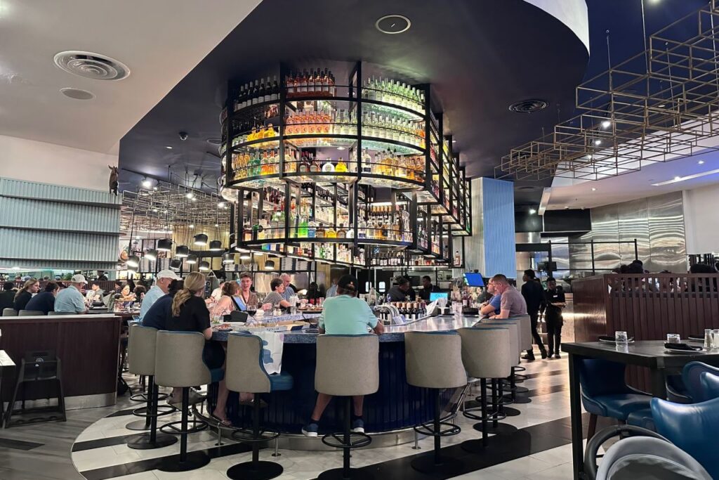 Busy restaurant interior at Baha Mar with a large circular bar stacked with liquor bottles serving as the centerpiece, surrounded by guests on bar stools.