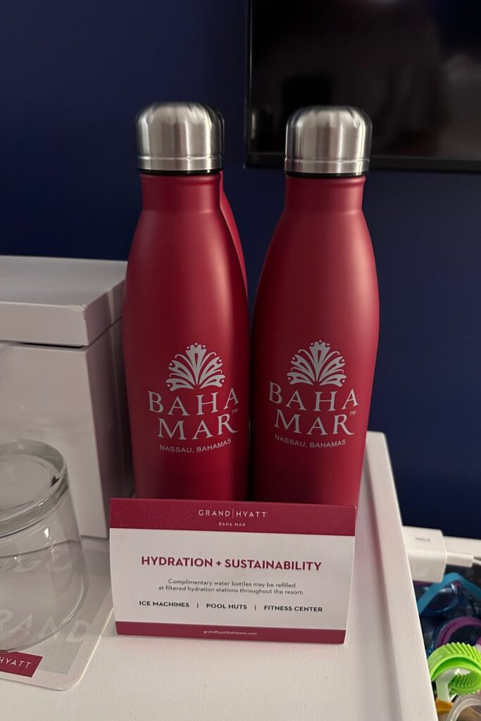 Two red Baha Mar branded reusable water bottles on a room desk alongside a card explaining the resort's hydration and sustainability program, at the Grand Hyatt Baha Mar.