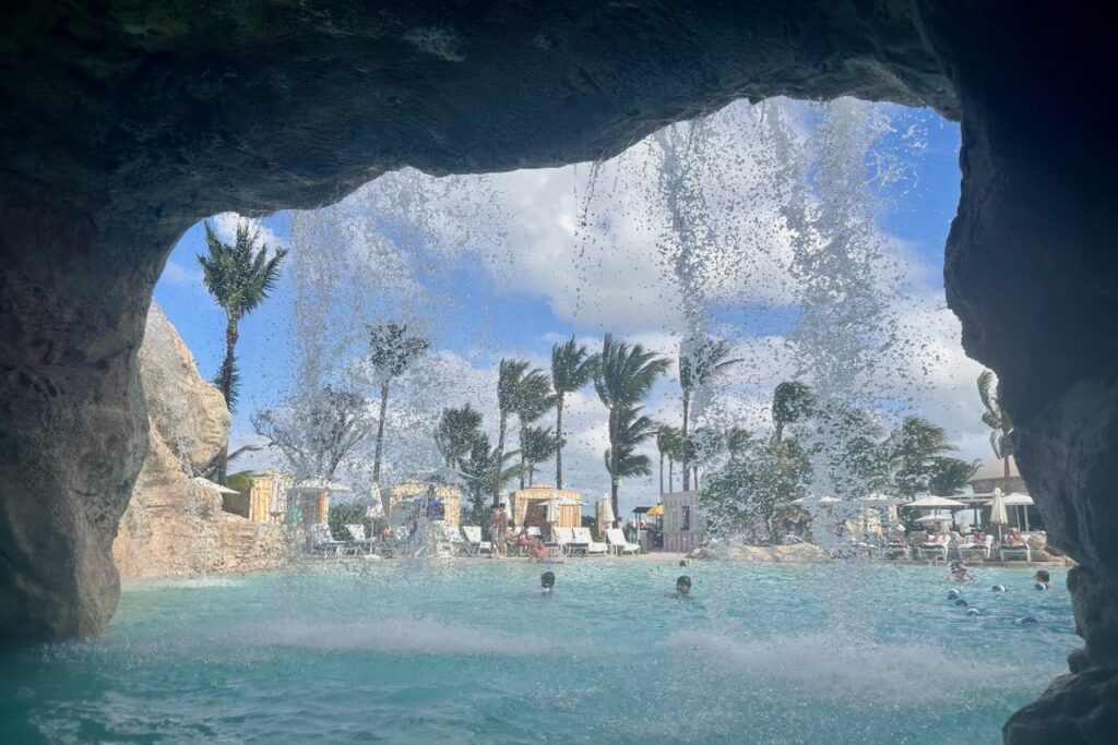 View from inside the grotto cave at the Dean's Blue Hole pool at Grand Hyatt Baha Mar, looking out through a waterfall curtain at the pool area and palm trees beyond.