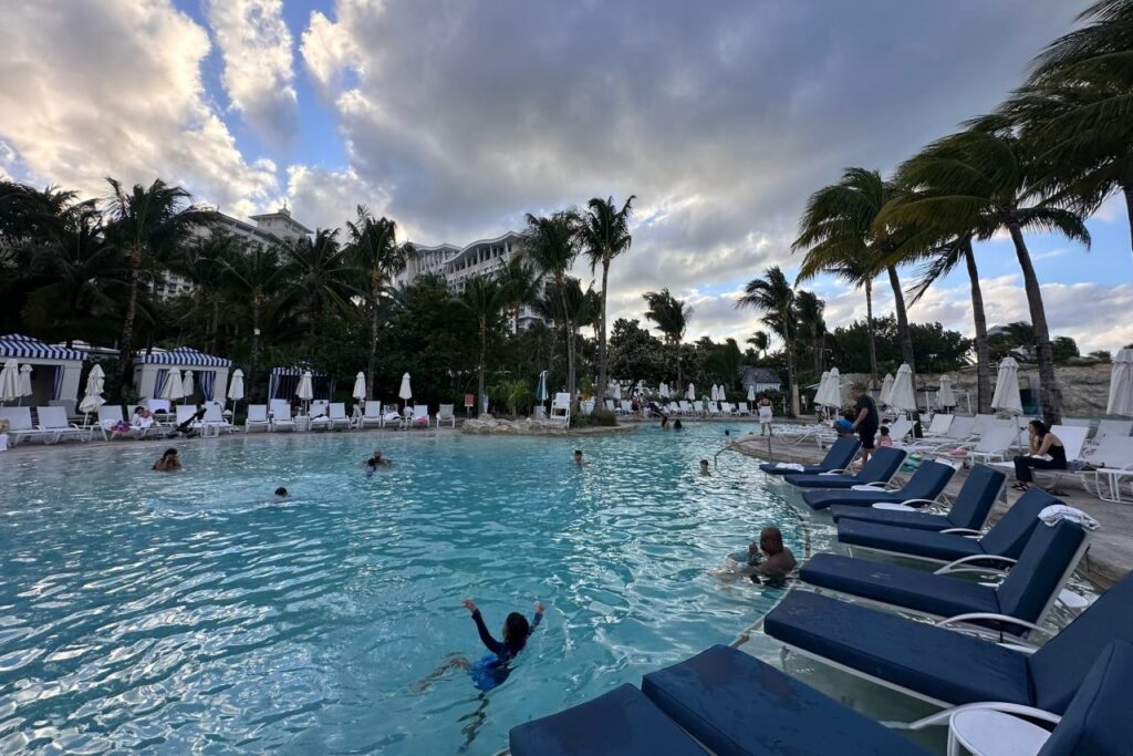 Guests swimming in one of the Grand Hyatt Baha Mar pools at dusk, with rock formations, a waterfall, and the resort tower visible in the background.
