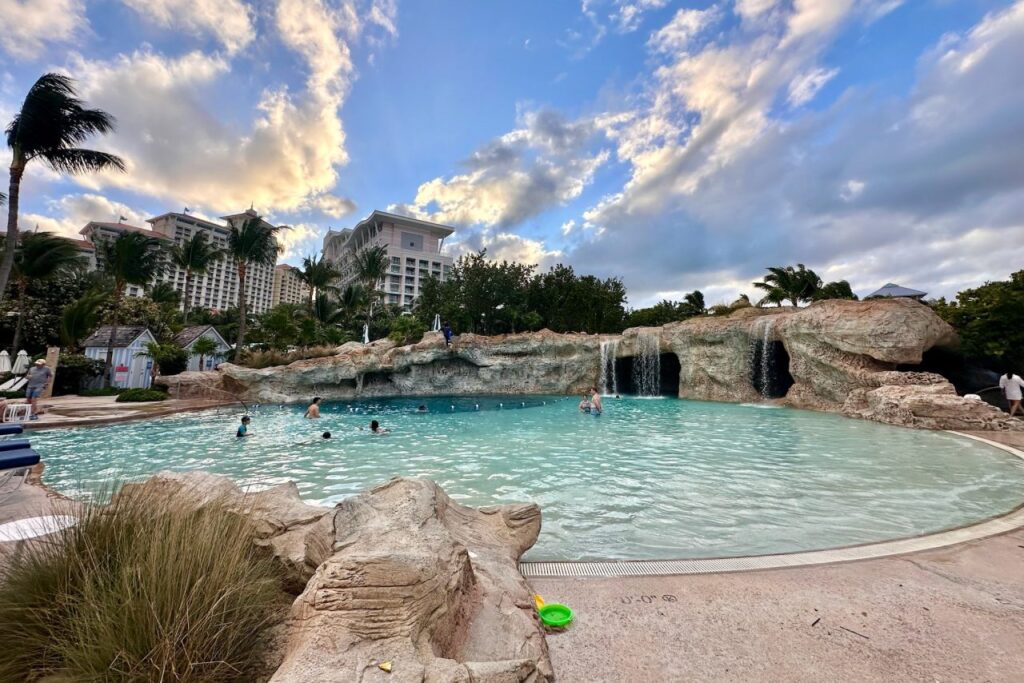 The Dean's Blue Hole pool at Grand Hyatt Baha Mar, featuring a rock waterfall, cave entrance, and guests swimming under a dramatic cloudy sky with the Rosewood tower in the background.