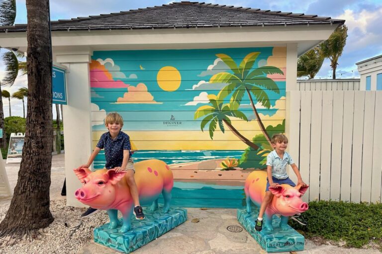Two young boys sitting on colorful pig sculptures in front of a vibrant Discover Bahamas mural at the Baha Mar resort, smiling at the camera.
