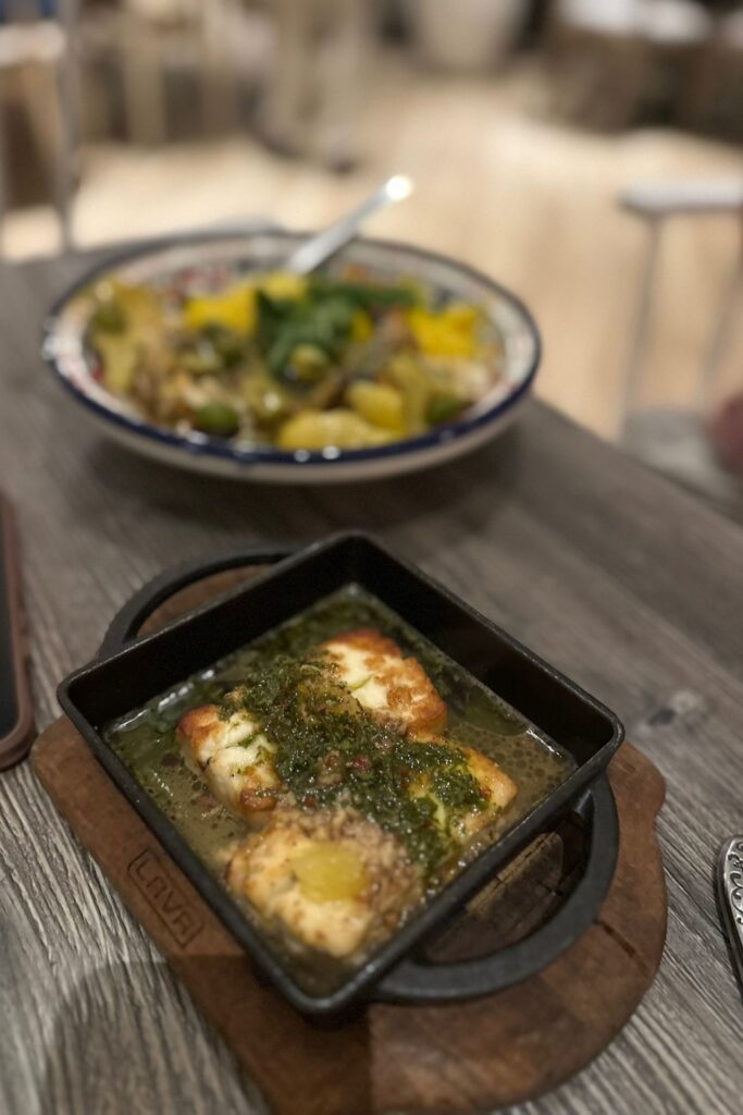 Seared halloumi in a cast iron skillet with herb sauce at a Baha Mar restaurant, with a colorful Moroccan-style dish visible in the background.