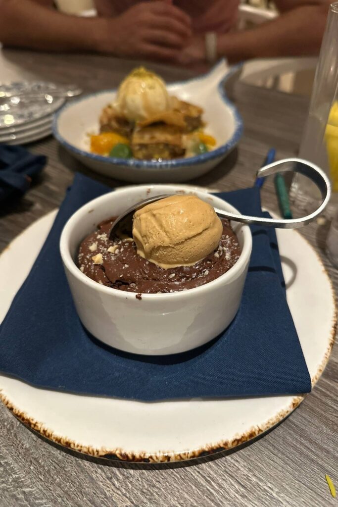 Warm chocolate brownie topped with a scoop of peanut butter ice cream in a white ramekin at a Baha Mar restaurant, with a baklava dessert visible in the background.