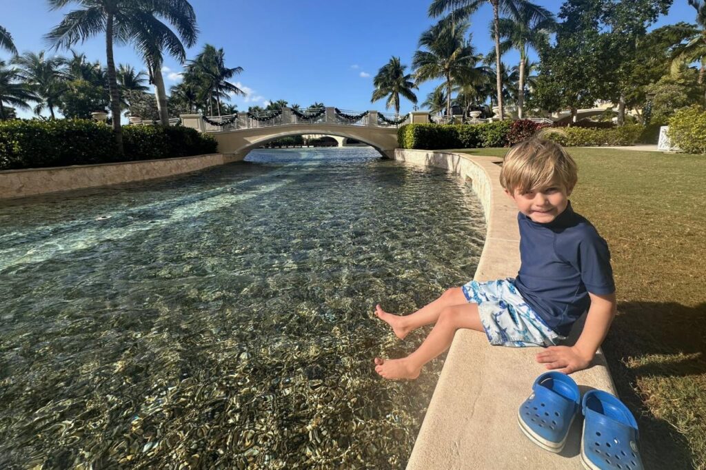 Young boy sitting barefoot on a low wall along a crystal-clear canal in the Grand Hyatt Baha Mar grounds, with an arched bridge and palm trees in the background.