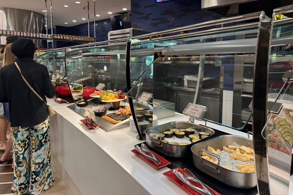 The Regatta breakfast buffet at Grand Hyatt Baha Mar, showing a long counter with hot dishes, shrimp, fruits, and pastries with a guest browsing the spread.
