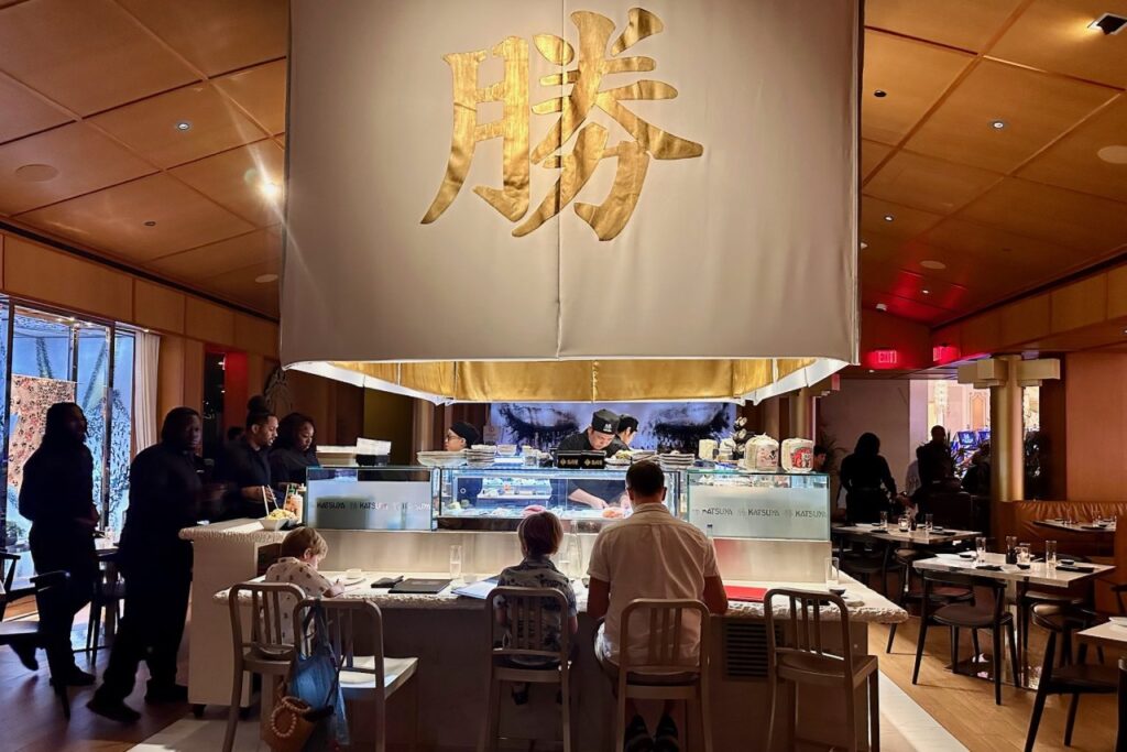 Interior of Katsuya restaurant at Grand Hyatt Baha Mar with a large gold calligraphy banner hanging above the sushi bar, where chefs are at work and families are seated.