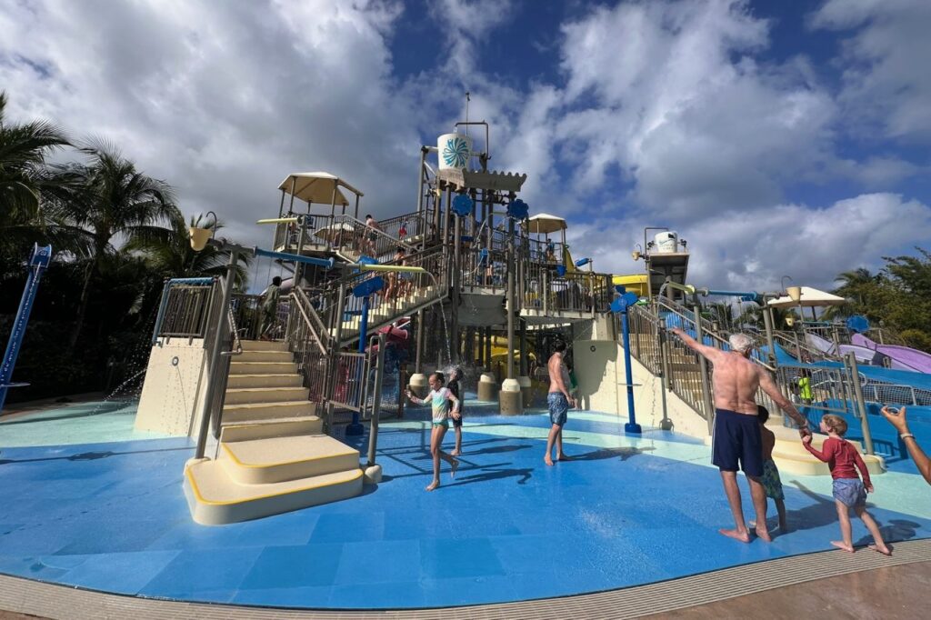 Families enjoying the kids' splash pad and multi-level water play structure at Baha Bay water park, Grand Hyatt Baha Mar, under a partly cloudy sky.