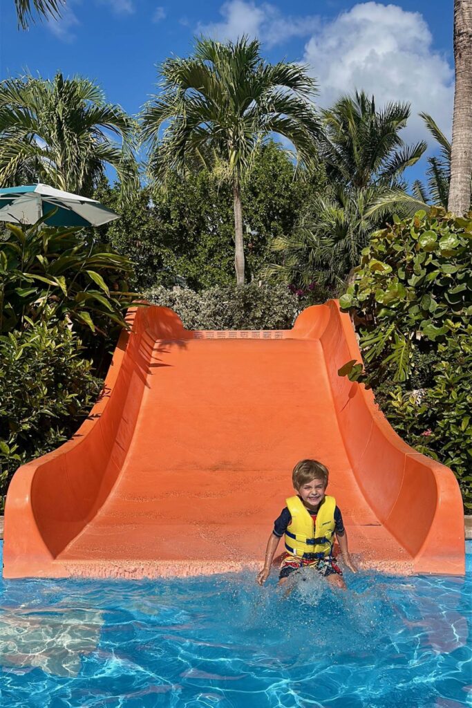 A young boy in a yellow life jacket landing at the bottom of an orange water slide at Baha Bay water park, Grand Hyatt Baha Mar, with a big smile on his face.
